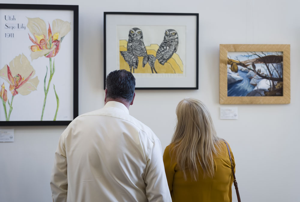 A man and a woman looking at a set of three paintings in the art gallery housed at UBTech's vernal campus. The first, on the left, is a painting of two sego lillies in white, orange, and gold. The middle painting is of two black and white owls perched on golden rock. The painting on the right is of a creek during winter time. 