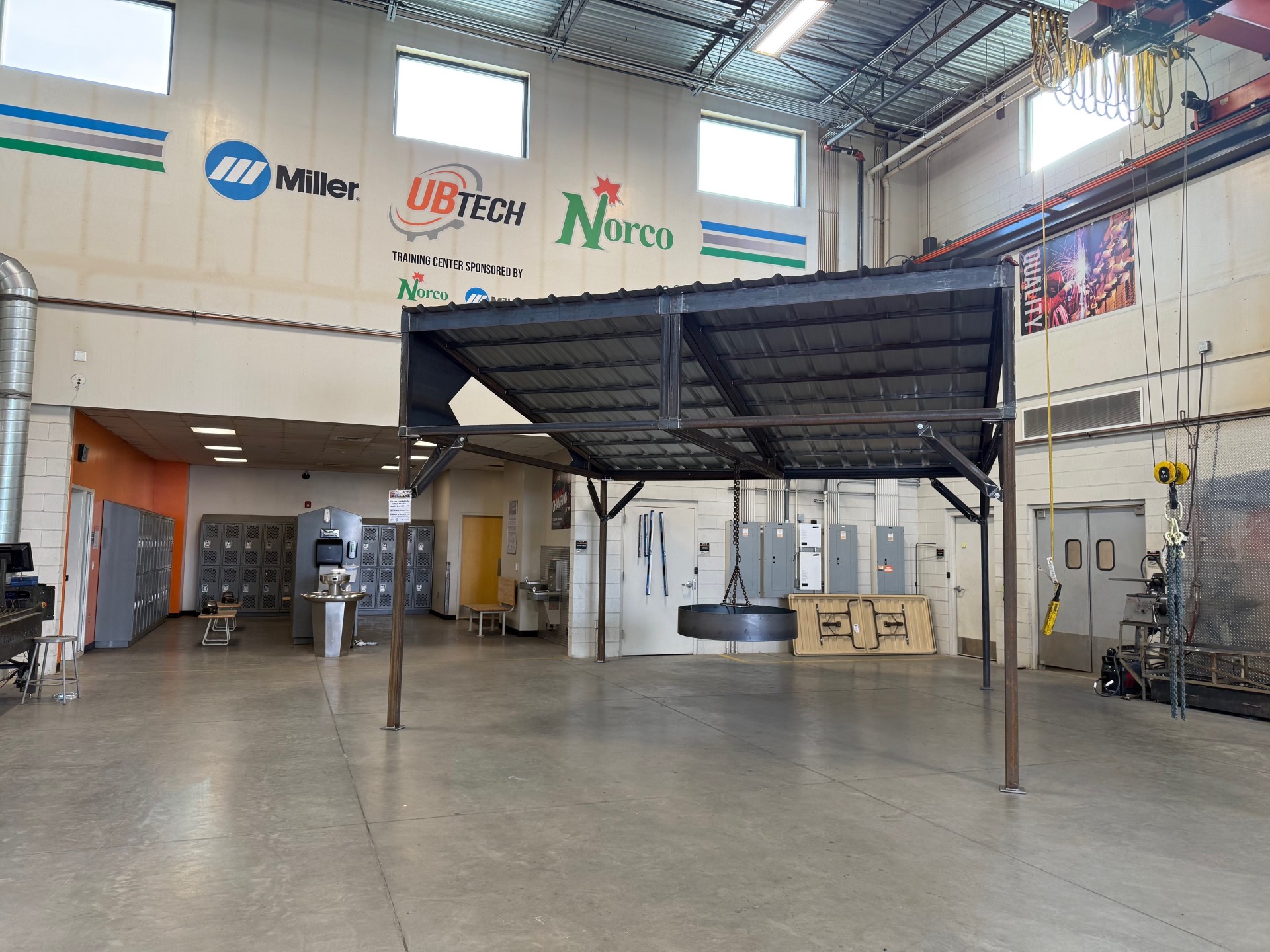 Gazebo with Firepit A metal, rectangular structure with close roof. It has support beams that hold the roof but no walls. It sits in a workshop. A circular firepit hangs from the roof