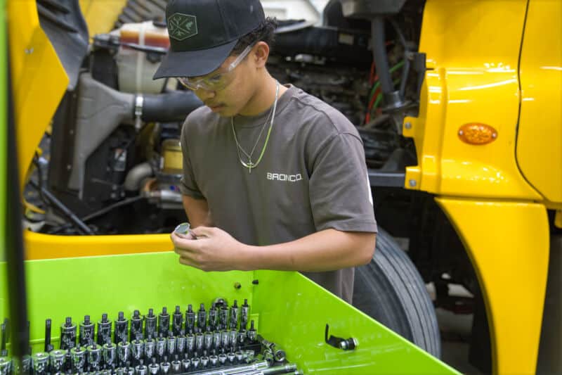 Male student looking into the bright lime green Carlyle tool box holding a socket standing in front of a yellow semi truck.
